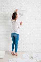 Female artist looking at her work in workshop. Barefoot woman standing at wall and hanging her pencil portrait on it, free space on white brick background. Art, talent, evaluating concept