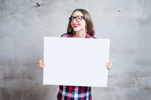 Young Woman Dressed Casually Holding White Board Or Empty Placard With Copyspace Standing On The Gray Wall Background.