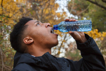 Young black man drinking water from a plastic bottle in the field on a colorful autumn day. 