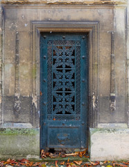 Old and weathered blue colored closed iron door in a stone wall of an old tomb / crypt at a graveyard. Artistic curvy flower ornaments and geometric forms, surrounded by rectangle frames / borders.