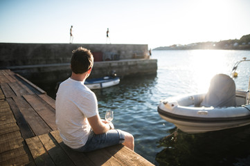 Young man sitting on wooden pier, holding glass of wine