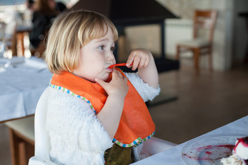 two years old child eating with orange plastic spoon and bib a piece of chocolate cake with vanilla ice cream at restaurant
