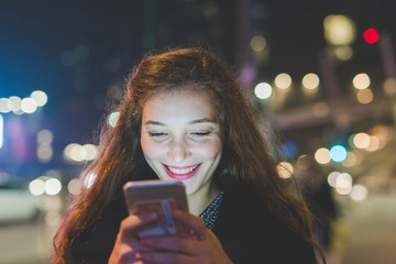 young beautiful caucasian long brown hair woman using smart phone hand hold outdoor in the city night, smiling, face illuminated screenlight - social network, technology, comunication concept