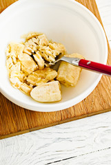 Wall Mural - Mashing banana for panckakes with red fork in white bowl, white wooden rustic background