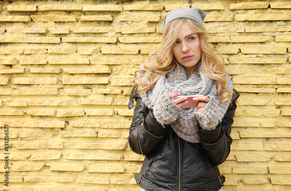 Young beautiful woman with smartphone standing on yellow brick wall background