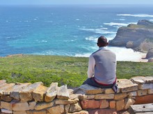 Contemplative African Young Man Sitting On The Rocks, Looking At Distance To The Blue Sea And Thinking About Something. The Cape Point In South Africa As A Background.