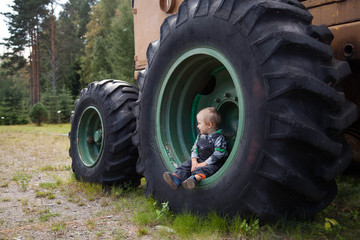 Little cute kid sitting in a huge wheel of the tractor. Wood mining machine.