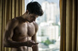 © theartofphoto - Sexy handsome young man standing shirtless holding a cell phone, in his bedroom next to window curtains