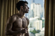 © theartofphoto - Sexy handsome young man standing shirtless holding a cell phone, in his bedroom next to window curtains