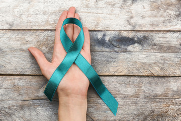 Poster - Female hand holding sea-green ribbon on wooden background. Ovarian cancer and gynecological disorders concept