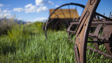 Farming Ranch Background With Barn And Rusty Farm Plow. Green Grass, Blue Sky And Wooden Barn. Shallow Depth Of Field With Focus On Rusty Plow Wheel.