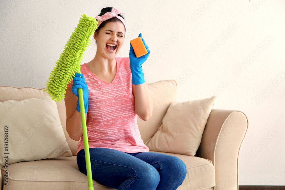 Happy young cleaner with dust mop and sponge sitting on sofa