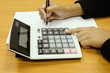 © Rapheephat - Office lady hand in a black suit holding a pen and pressing button on a calculator with a plain white paper on a wood desk background