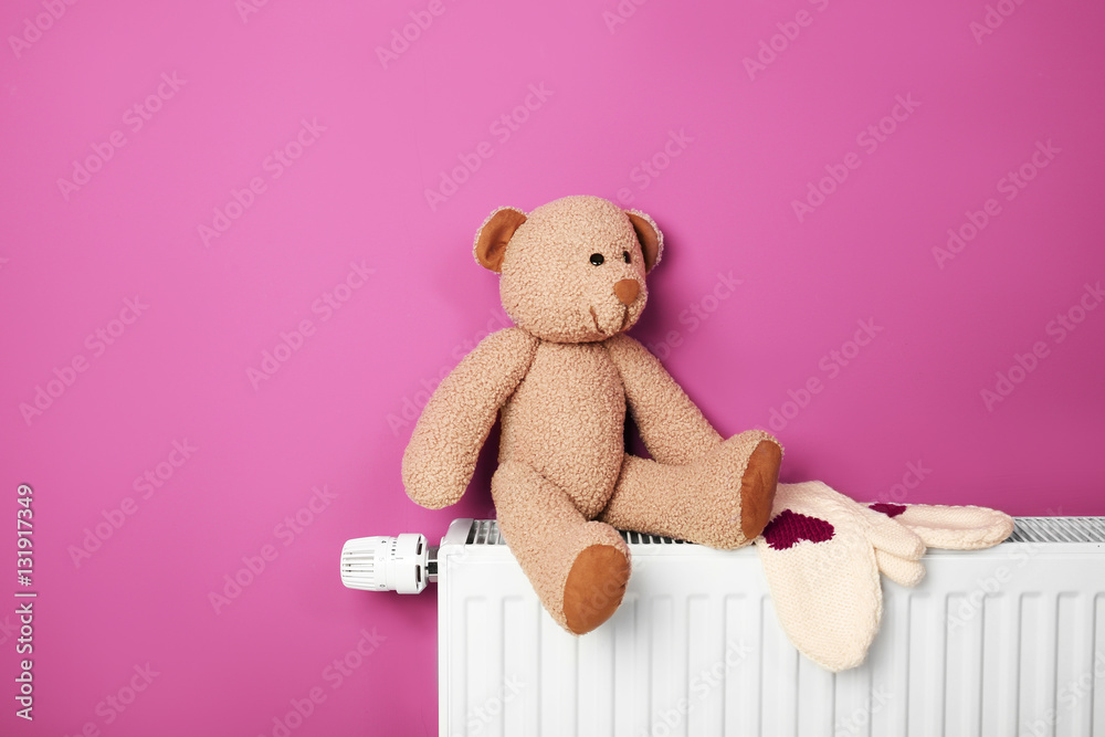 Teddy bear with mittens on heating radiator near pink wall