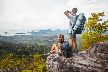 Poster - Couple of tourist with backpacks relaxing on top of a mountain and enjoying the view of valley