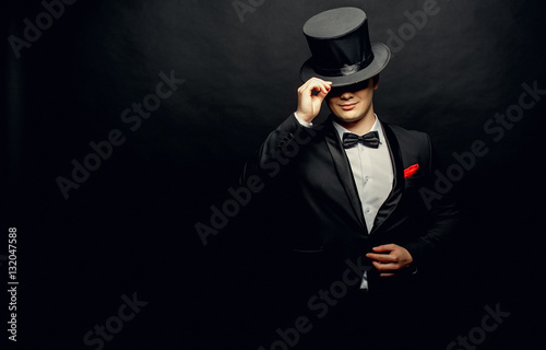 Fotomural A magician in a black suit holding an empty top hat and magic wand isolated on b