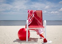 Horizontal Image Of A Red Beach Chair With A Red And White Towel Hanging Over The Back And A Big Red Beach Ball Lying On The Sandy Beach With The Lake In The Background In The Summer Time.