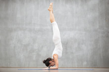 Portrait Of Beautiful Young Woman Wearing White Sportswear Working Out Against Grey Wall, Doing Yoga Or Pilates Exercise. Handstand Feathered Peacock Pose, Pincha Mayurasana. Full Length Shot