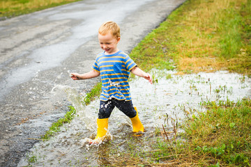 Smiling funny toddler boy in yellow rubber boots, playing with water after the rain. He is running on the wetroad. 