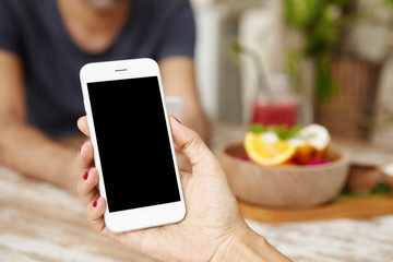 People, technology and lifestyle concept. Close up shot of female hand holding smart phone, browsing internet and messaging friends online via social networks sitting in blurred interior of cafe