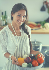 Wall Mural - Smiling young woman holding vegetables standing in kitchen