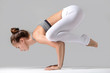 © fizkes - Young attractive woman practicing yoga, standing in Crane exercise, Bakasana pose, working out wearing sportswear, white pants, gray top, indoor full length, isolated against grey studio background