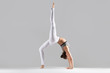 © fizkes - Young attractive woman practicing yoga, standing in Bridge exercise, One legged Wheel pose, working out wearing sportswear, white pants, indoor full length, isolated against grey studio background