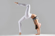 © fizkes - Young attractive woman practicing yoga, standing in Bridge exercise, One legged Wheel pose, working out wearing sportswear, bra, indoor full length, isolated against grey studio background