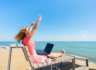 I'm the winner - young woman sitting with laptop, coffee cup and smartphone on the beach.