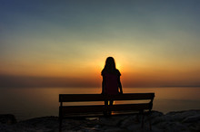 The Silhouette Of A Lonely Girl Sitting On A Bench On The Top Of The Edge Of A Cliff And Watching The Sunset