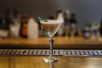 A drink in a cocktail (martini) glass with dressing and mint leaf on the top, on the bar stand. Small depth of field. Selective focus. Film grain effect.