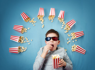 Child lying on blue background in 3d glasses and watching tv. Boy eating popcorn