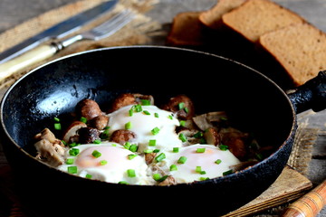 Wall Mural - Fried eggs in pan. Eggs fried with mushrooms and green onions. Brown bread slices, fork, knife on a wooden table. Quick vegetarian breakfast recipe with eggs. Vintage style. Closeup