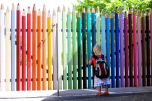 Happy Little Girl Going To School. Cute Preschooler Kid With Backpack Enjoying First School Day. Little Student Standing In Front Of The Colorful Pencil Shape Fence Looking At The Schoolyard. 