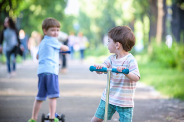 Two cute boys, compete in riding scooters, outdoor in the park, summertime.