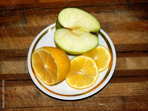 On A Wooden Chopping Board In A White Saucer With Golden Rim Is Half Green Apple And Lemon And Two Slices Of Lemon Cut On A Horizontal Surface In Front View Stock