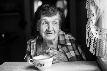 Elderly woman, drinking tea sitting at the table in the house. Black-and-white portrait.