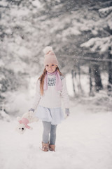 Wall Mural - Cute smiling kid girl 4-5 year old holding teddy bear walking in snow in park. Looking at camera. Childhood. Wearing knitted hat, sweater and skirt outdoors.