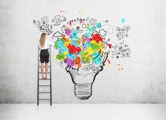 Wall Mural - Rear view of a blond woman standing on a ladder and drawing a large and colorful light bulb sketch on a concrete wall.