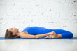 © fizkes - Young attractive woman practicing yoga, lying in Reclining Hero exercise, Supta Virasana pose, working out wearing sportswear, blue suit, indoor full length, white studio background