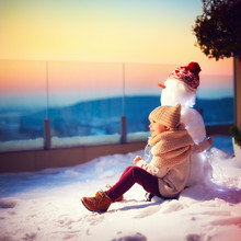 Happy Little Kid And His Friend Snowman Watching The Sun Goes Down Sitting In Snow On Rooftop Terrace In One Winter Evening