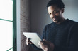 Selective focus.Smiling bearded African man reading news digital tablet while standing near the window in his modern apartment.Concept of young business people working at home.Blurred background.
