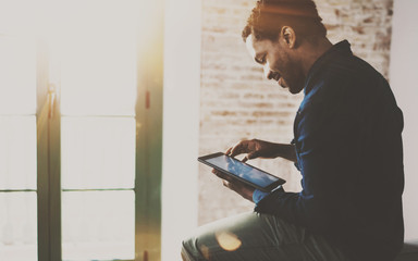 Musing young African American freelancer touching display of digital tablet while working in new project at home.Black businessman looking for information on Internet.Blurred,color filter,flares.