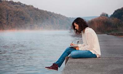 girl writing diary by the lake in the early cold morning.