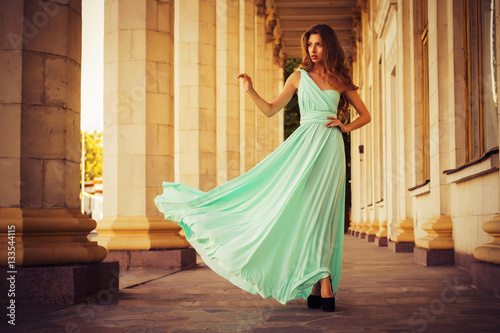 Foto Beautiful blonde with a long curly hair in a long evening dress in motion outdoors near retro vintage building in summer sunset