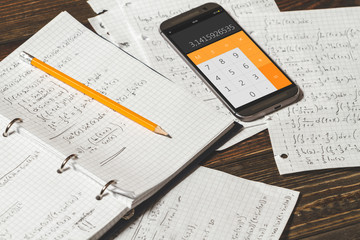 Solving mathematical problems in a notebook .  Phone with calculator app on wooden desk.