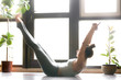 Young attractive woman practicing yoga, stretching in Paripurna Navasana pose, working out, wearing sportswear, grey pants, bra, indoor full length, home interior background, near the floor window