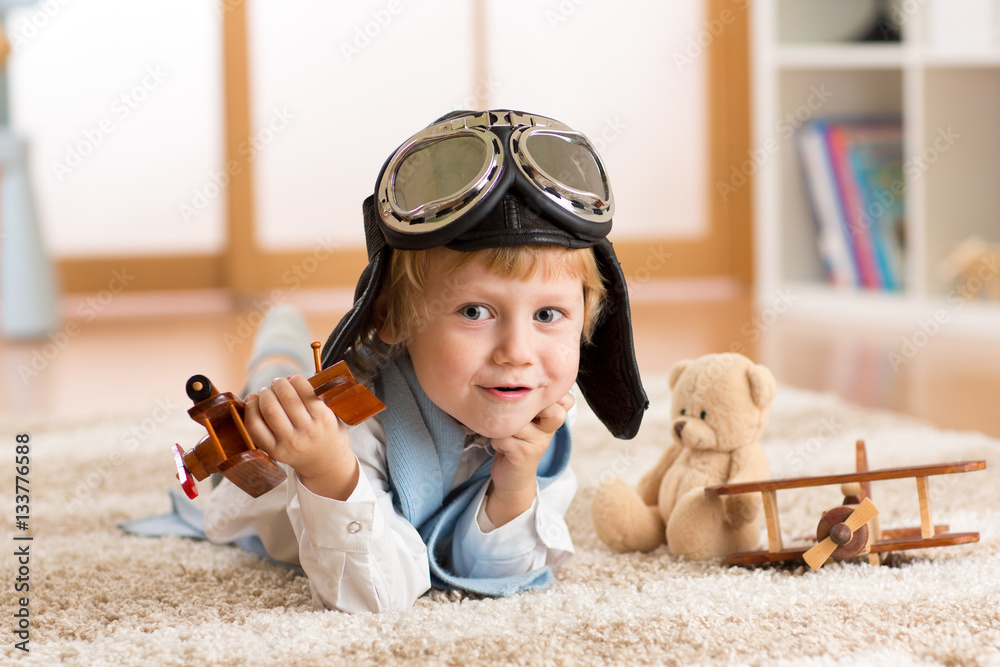 Child weared pilot or aviator plays with a toy airplane at home in nursery room. Concept of dreams and travels.