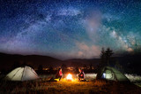 Fototapeta  - Romantic couple sitting at a campfire near tents in the night under incredibly beautiful starry sky and Milky way. Night camping