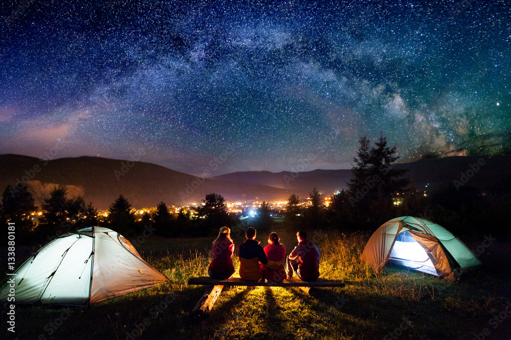 Friends hikers sitting on a bench made of logs and watching fire together beside camp and tents in the night. On the background beautiful starry sky, mountains and luminous town. Rear view - obrazy, fototapety, plakaty 